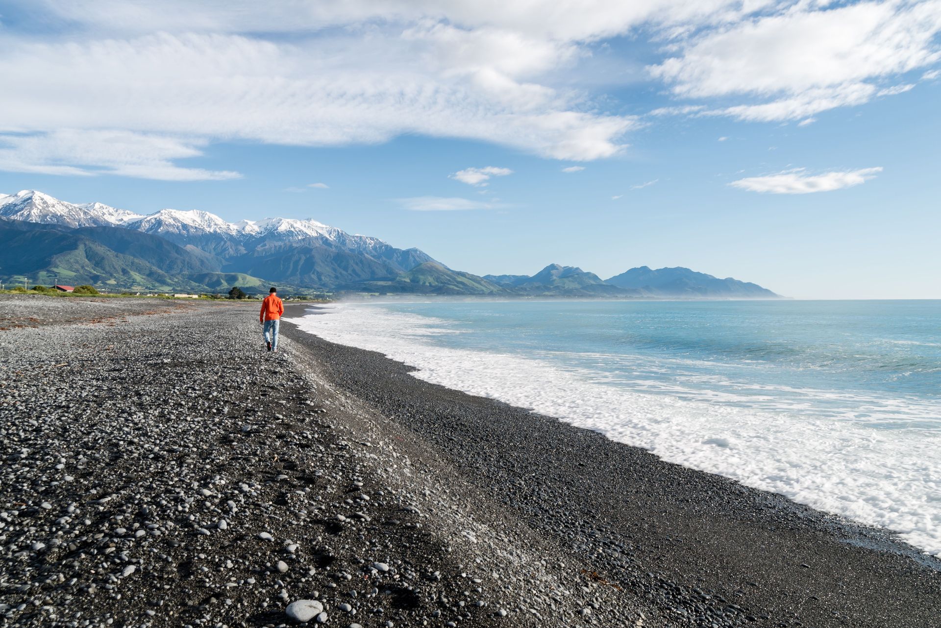 Un homme marche le long d'une plage rocheuse avec une vue imprenable sur des montagnes enneigées et l'océan.
