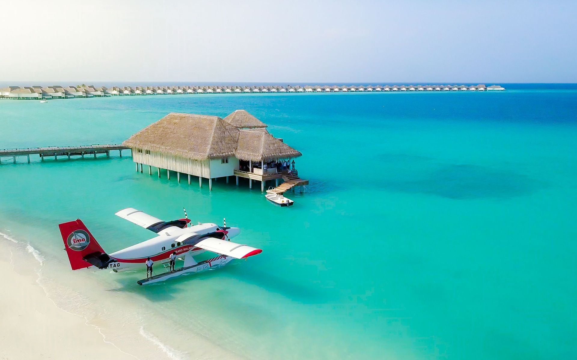 Une vue aérienne à couper le souffle de bungalows sur l'eau dans une mer turquoise avec un hydravion sur une plage de sable.