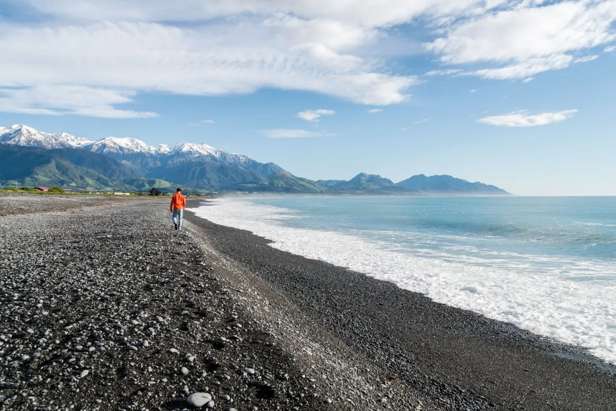 Un homme marche le long d'une plage rocheuse avec une vue imprenable sur des montagnes enneigées et l'océan.